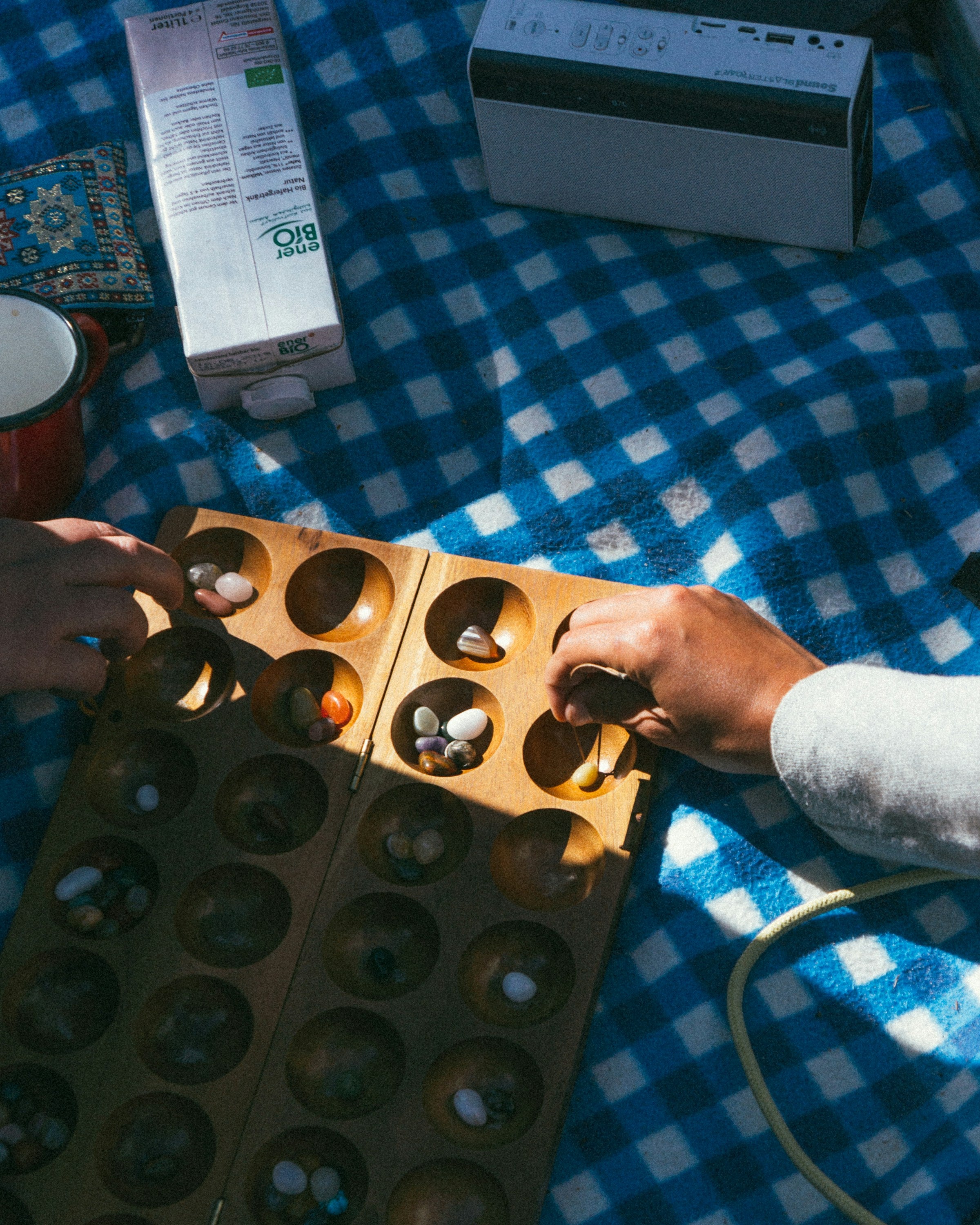 Two hands playing a game of Mancala on a blue checkered tablecloth.