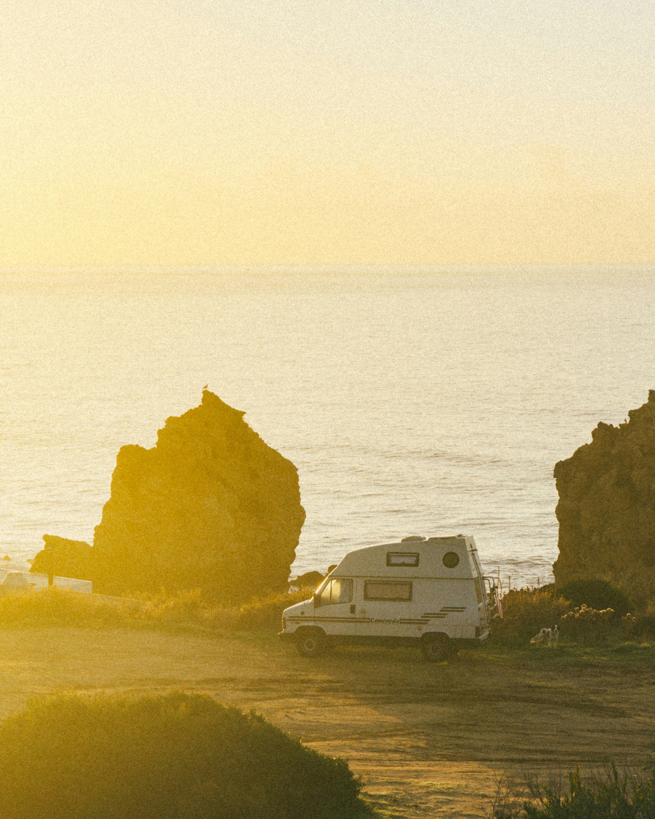 Van parked on a grassy area near the ocean with a sunset or sunrise in the background.